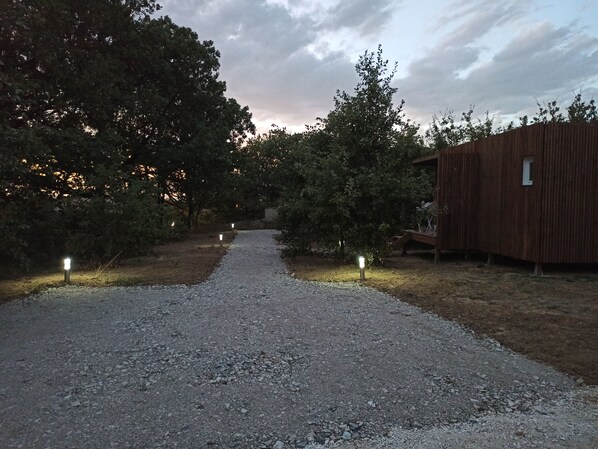 Un camí de grava que condueix a una construcció de fusta amb un petit finestral, envoltada d'arbres i il·luminada per llums al terra.