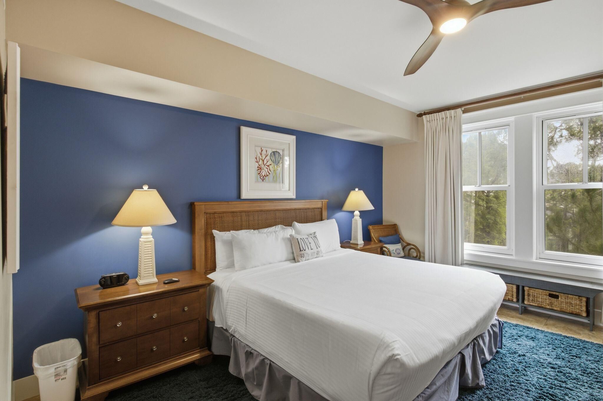 Une chambre à coucher comprenant un lit, deux tables de chevet, un ventilateur de plafond, une fenêtre avec des rideaux et un tableau accroché au mur.