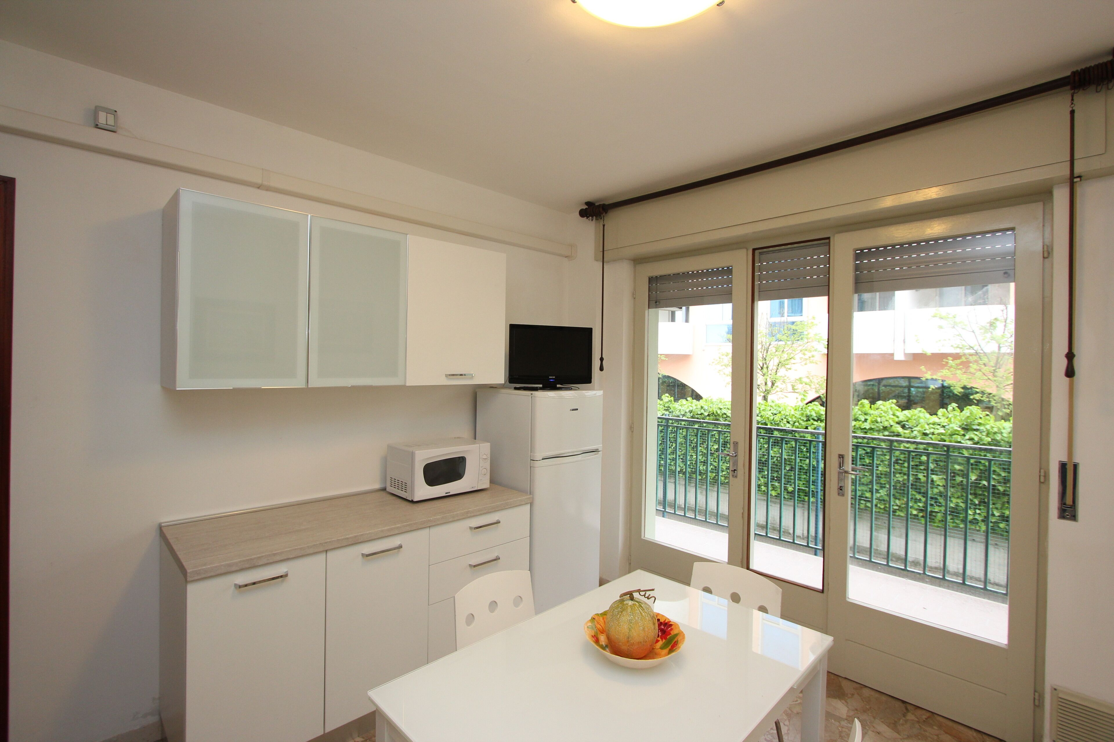 A compact kitchen with white cabinets, a microwave, and a small refrigerator. A dining table with chairs is positioned near a sliding glass door that opens to a balcony with a view of greenery.