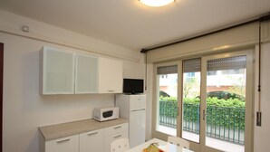 Une cuisine compacte avec des meubles blancs, un four à micro-ondes et un petit réfrigérateur. Une table à manger avec des chaises est placée près d’une porte-fenêtre coulissante qui donne sur un balcon offrant une vue sur la verdure.