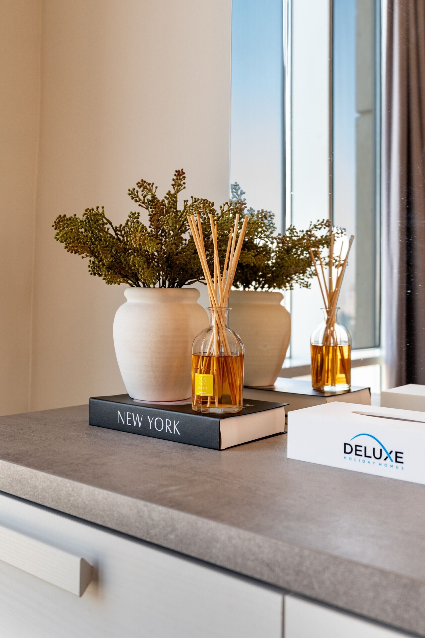 Una encimera de cocina moderna con una planta en maceta, dos botellas de vidrio con cañas de junco, un libro titulado 'NEW YORK' y un cartel de 'DELUXE HOLIDAY HOMES'.