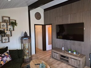A living room with a wooden floor, a TV mounted on a wall, a dark-colored sofa, and a decorative lantern. - Freistehender Ferienbungalow im Fachwerkstil (Butjadingen)