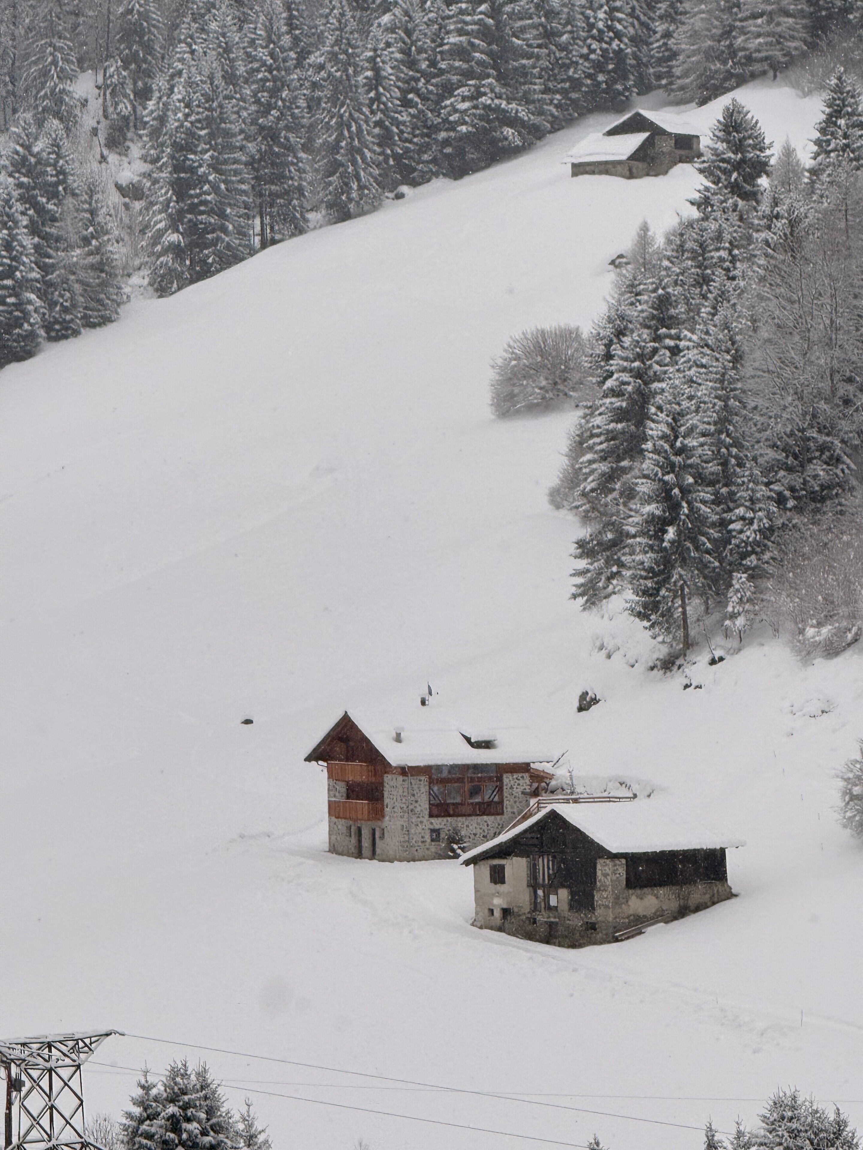 Une montagne enneigée, avec deux bâtiments : l’un avec un toit en bois et l’autre avec un toit foncé, le tout entouré d’arbres chargés de neige.