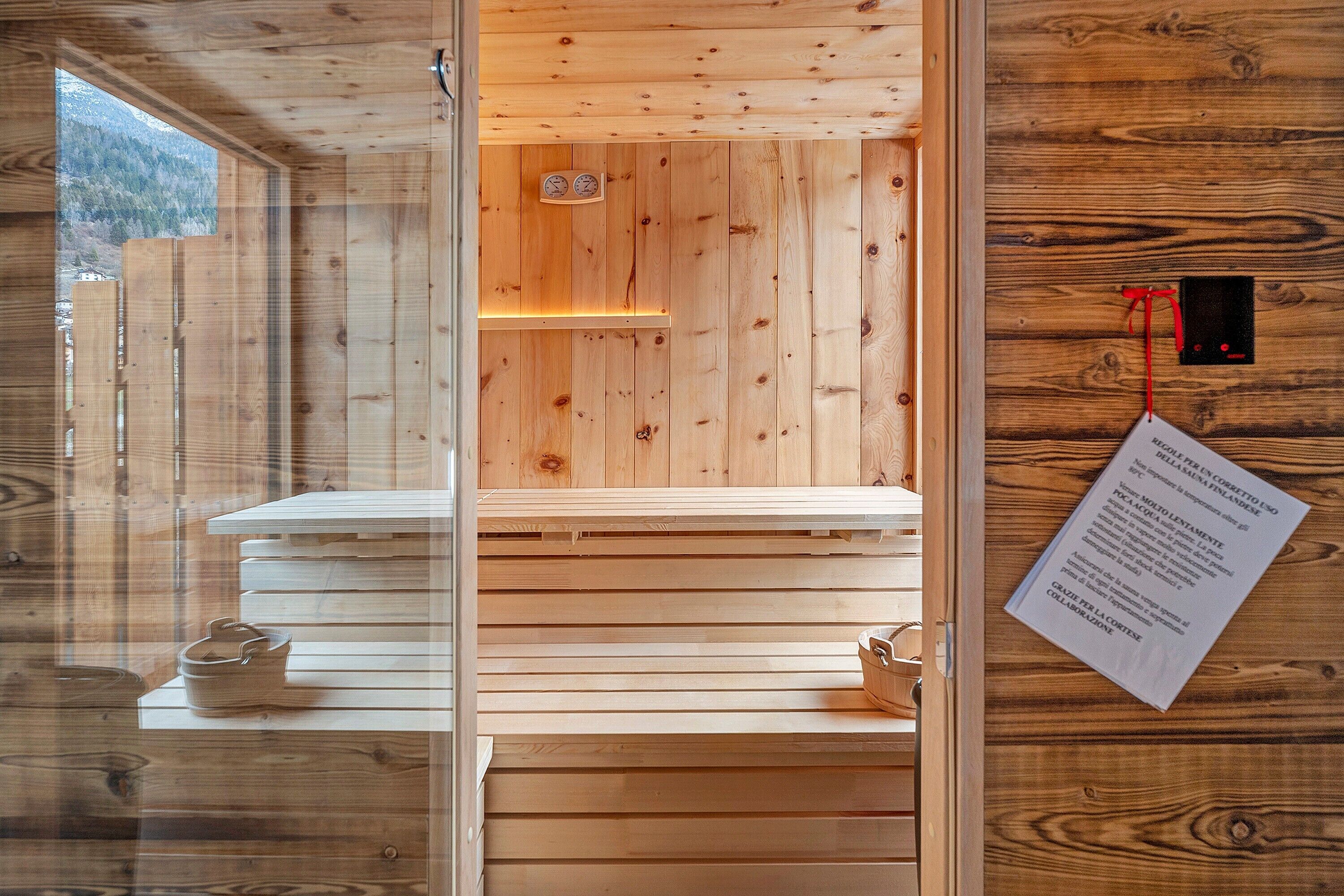 A wooden cabin interior with a sauna, wooden benches, a sauna door, and a view of the outdoors.