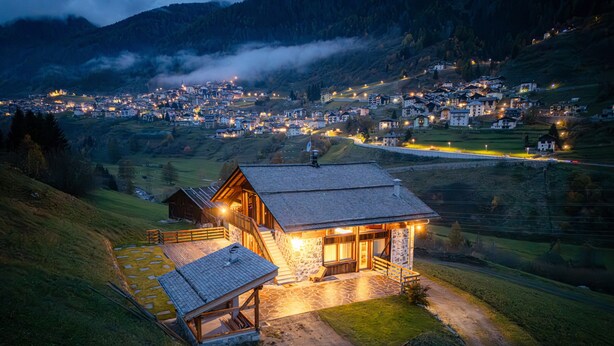 Una baita di montagna con facciata in pietra, un patio in legno e vista su una valle con case.