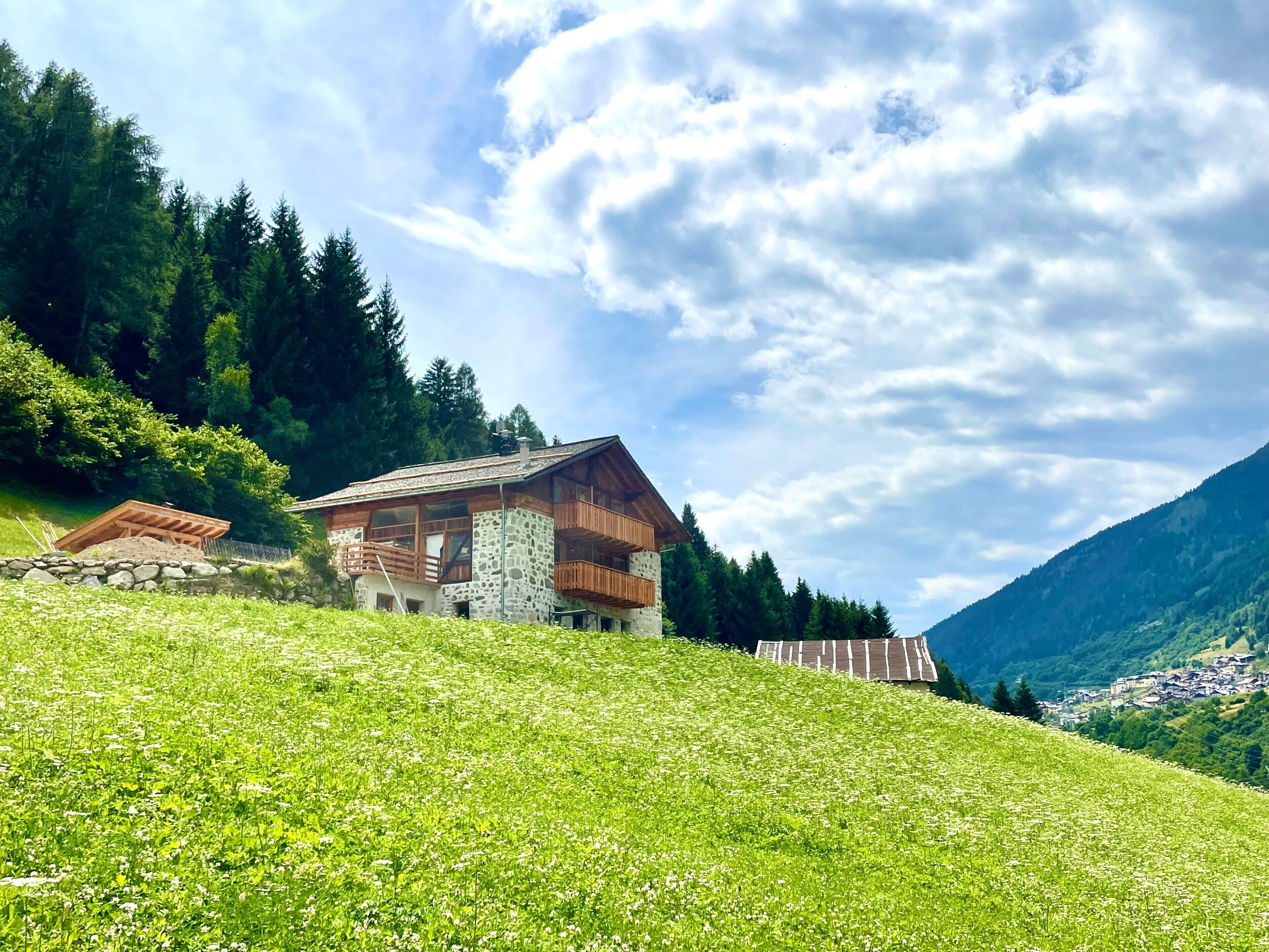 Una baita in pietra con balconi in legno, immersa nel verde e con montagne sullo sfondo.
