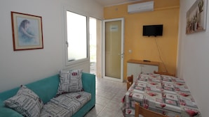 A small, well-lit room with a teal sofa, a dining table with a patterned tablecloth, a television mounted on the wall, and a door leading outside. - Rosolina Mare flat near beach (Rosolina Mare)