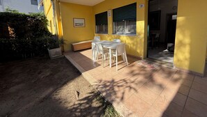 Un edificio amarillo con un patio que incluye una mesa, sillas, un banco y un pequeño jardín con plantas.