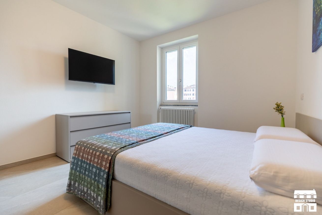 Ein Schlafzimmer mit einem Bett, einem flachbildfernseher an der Wand, einem Fenster mit Blick auf Gebäude und einem Heizkörper unterhalb des Fensters.
