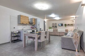 A modern kitchen with wooden cabinets, a dining area with a table and chairs, a grey sofa, and a TV mounted on the wall. - Villa Olive Groove Itria Valley (Puglia)