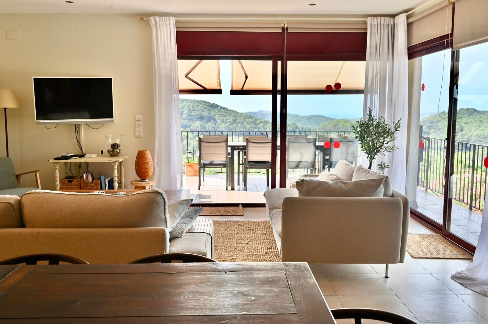 A modern living room with a flat-screen TV, a wooden table, and a sofa. There is a balcony with a view of the hills.