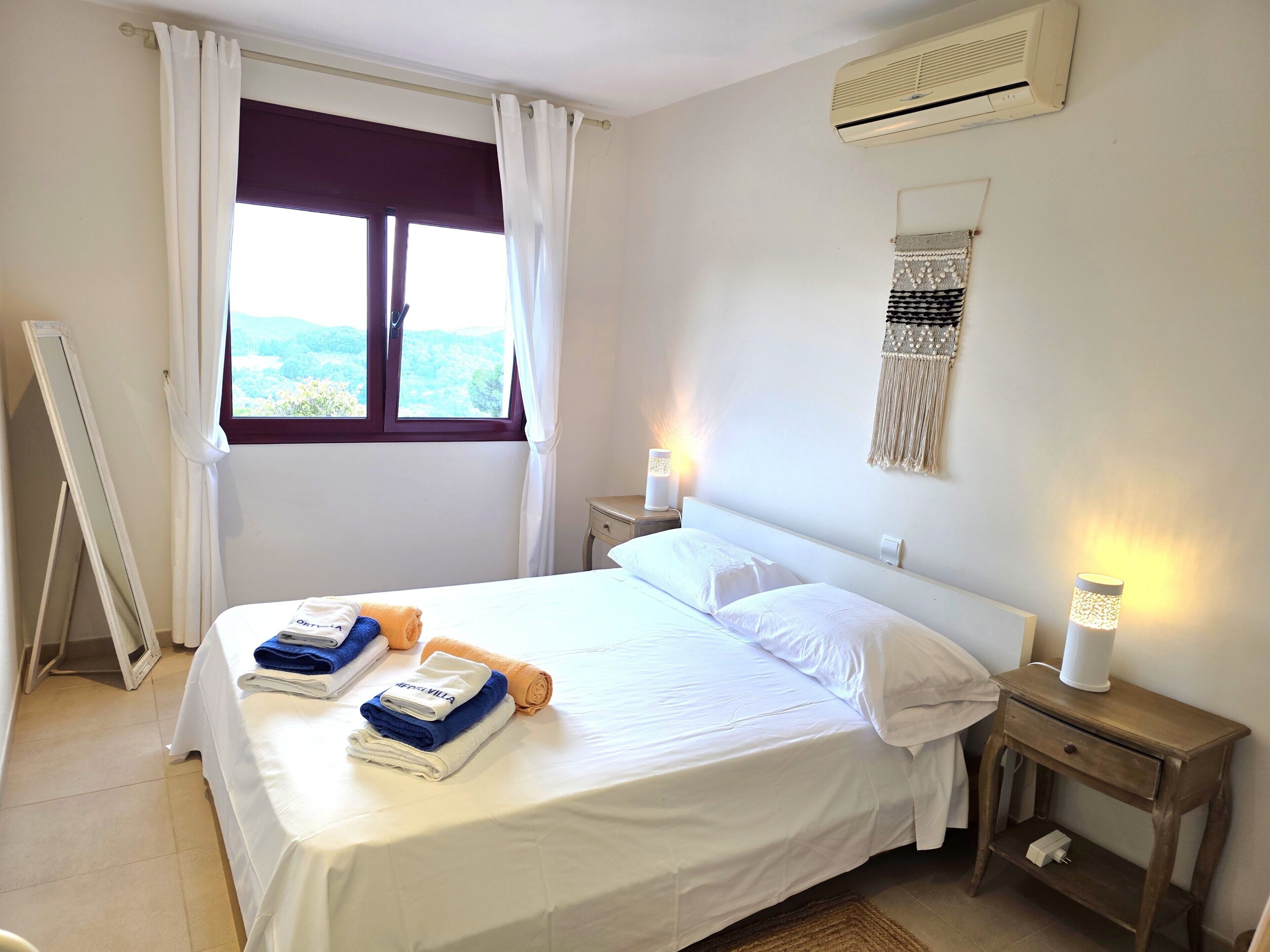 A hotel room with a bed, two bedside tables, a wall-mounted air conditioner, a window with curtains, and a view of the outdoors.