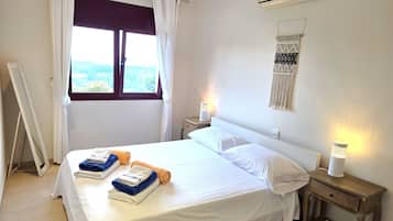 A hotel room with a bed, two bedside tables, a wall-mounted air conditioner, a window with curtains, and a view of the outdoors.