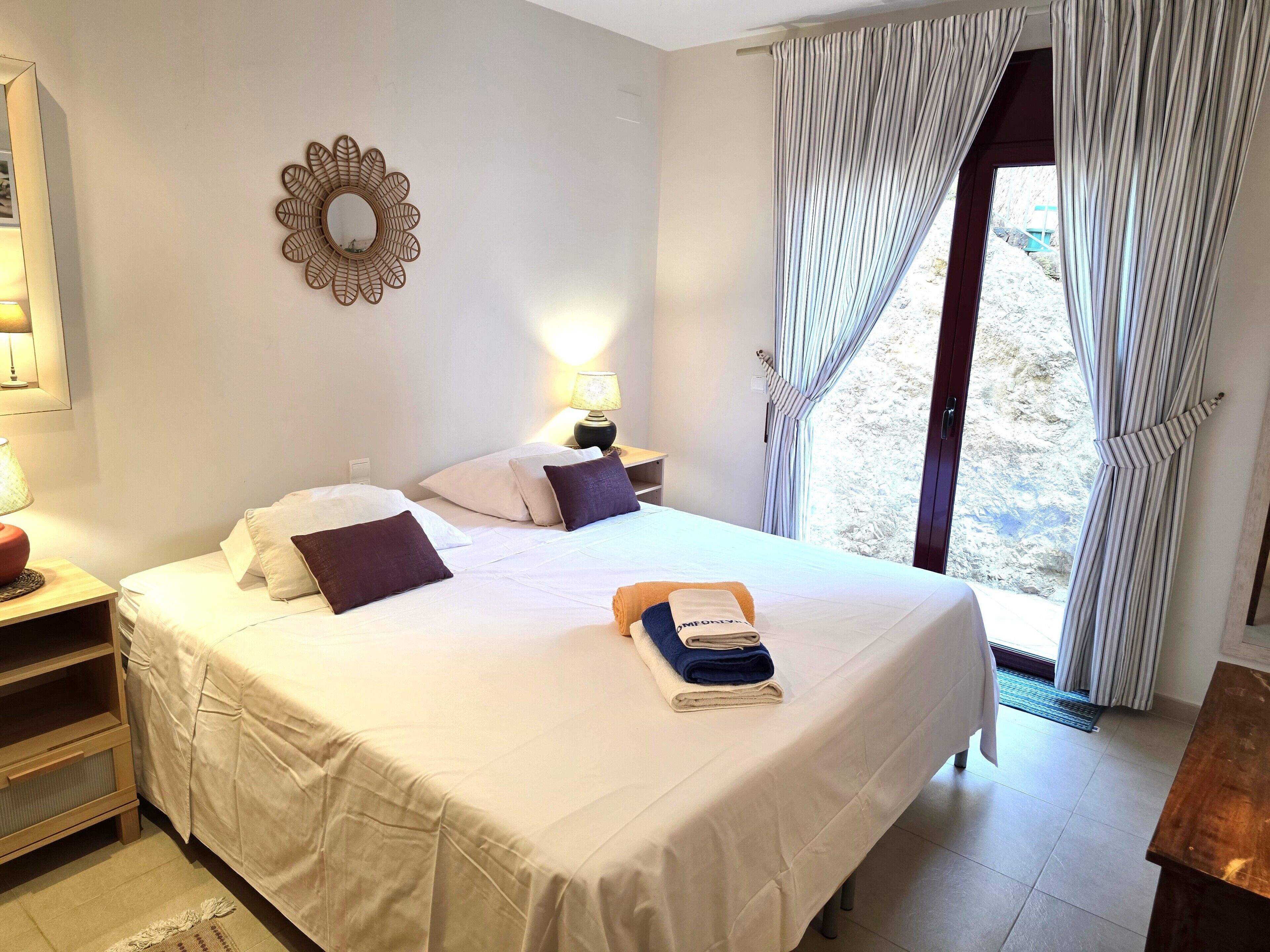 Una cama bien hecha con almohadas, una lámpara de noche y un ventanal con cortinas.