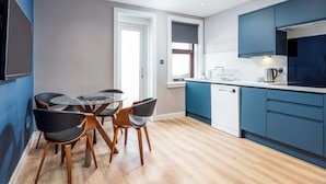Une cuisine moderne avec des meubles bleus, un coin repas avec une table en verre et des chaises en bois, et une vue sur la ville par une fenêtre.
