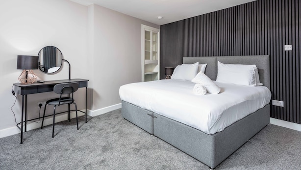 Une chambre à coucher moderne avec un lit gris, un bureau avec une chaise, un miroir rond et un luminaire au plafond.