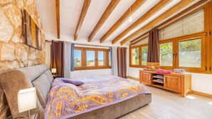 Une chambre avec un grand lit, des poutres en bois, un mur en pierre et de grandes fenêtres.