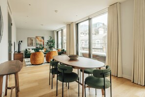Un salon moderne avec une table à manger en bois, des chaises recouvertes d’un tissu vert, un canapé vert et une grande fenêtre avec des rideaux.