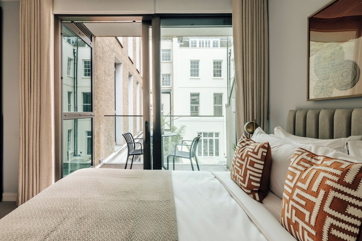 A hotel room with a bed, a balcony with a chair, and a view of buildings.