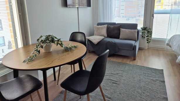 A modern living room with a round wooden table, black chairs, a grey sofa with cushions, and a large window with blinds. - M&J Apartments Harbour (Jyväskylä)