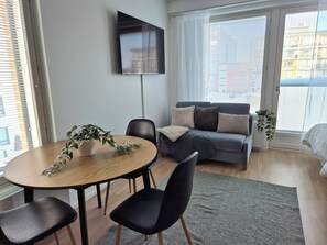 A modern living room with a round wooden table, black chairs, a grey sofa with cushions, and a large window with blinds. - M&J Apartments Harbour (Jyväskylä)