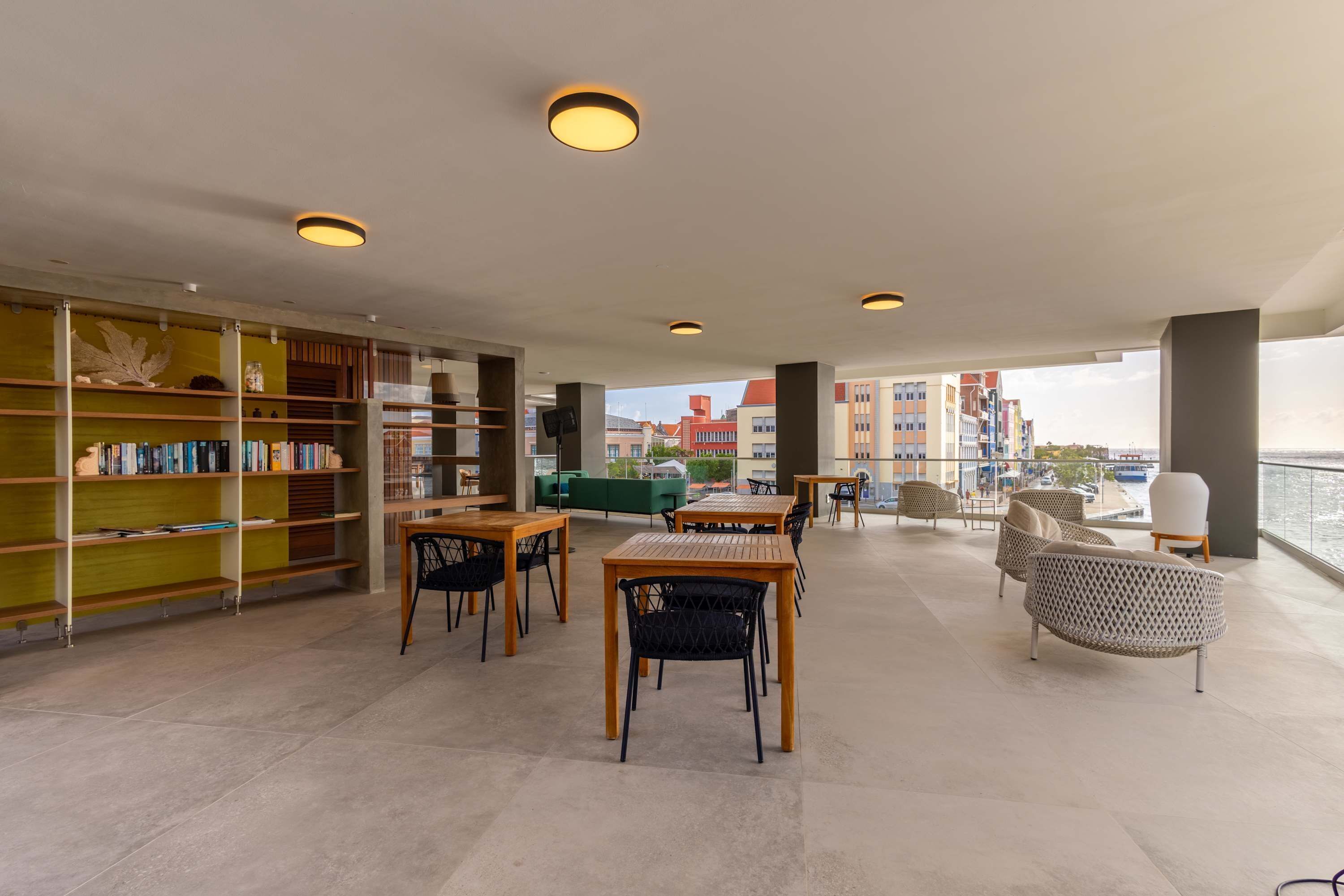 A modern library with bookshelves, tables, and chairs, overlooking a waterfront.