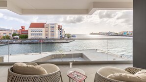 A balcony with wicker furniture, a glass table, and a view of a waterfront with buildings and a dock. - The Wharf 5A in Willemstad (Willemstad)
