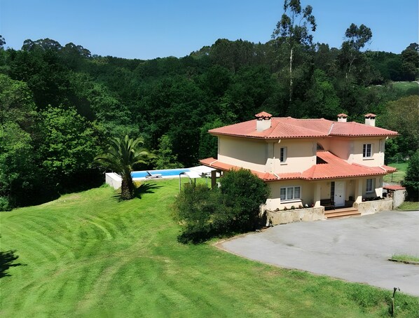 Une maison à deux étages avec une piscine, entourée d’une végétation luxuriante et de collines.