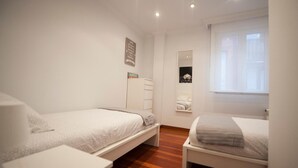 Une chambre avec deux lits simples, une armoire blanche, un miroir et une fenêtre avec des stores.