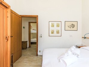 Un dormitorio con un armario de madera, una cama con ropa blanca, un pasillo que lleva a un baño y dos cuadros en la pared.
