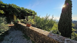 En mur av natursten som fungerar som stödmur, med krukväxter, en pergola och ett högt cypressträd.