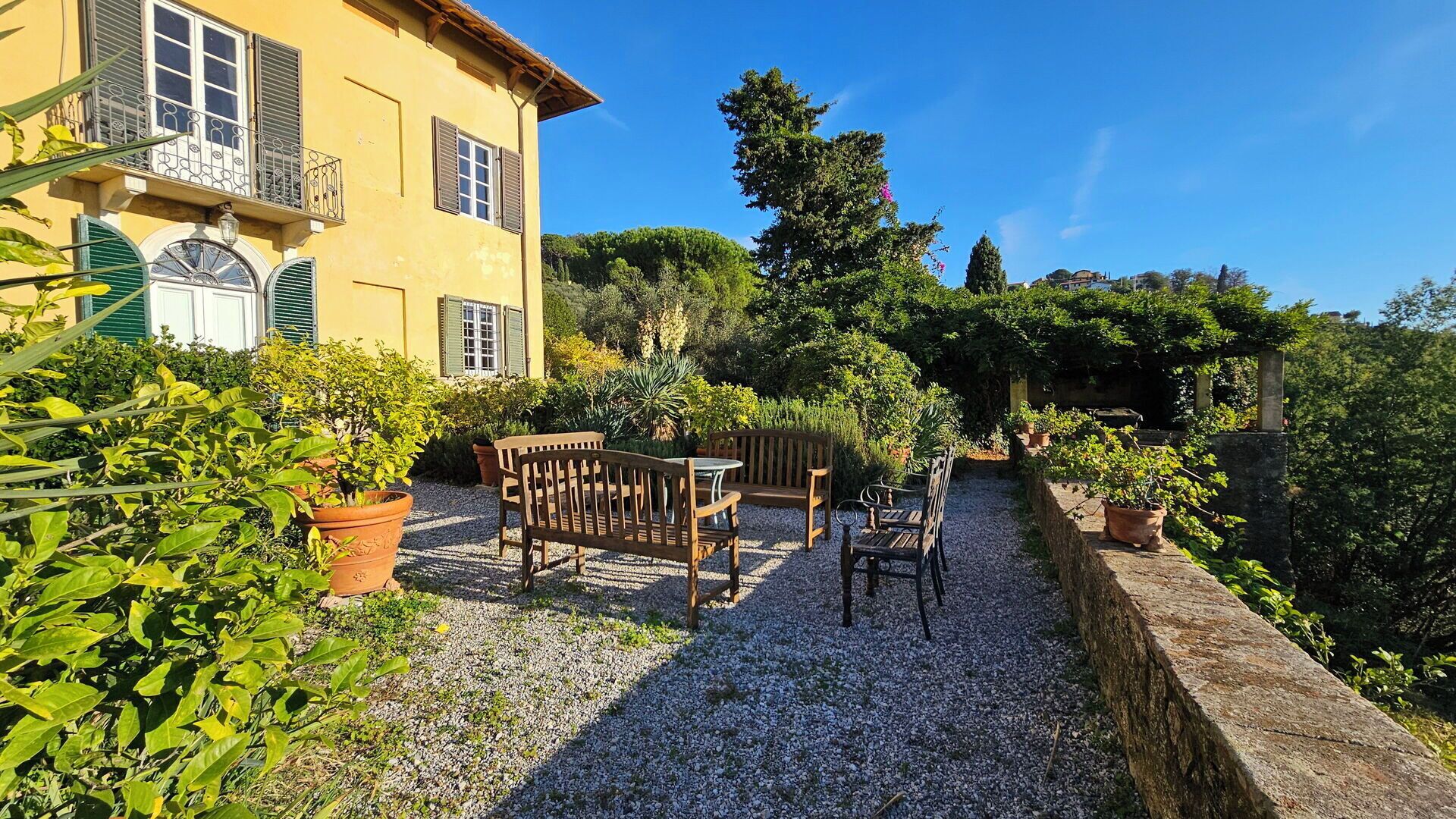 Una villa amarilla con un patio de grava, mobiliario exterior y plantas en macetas.