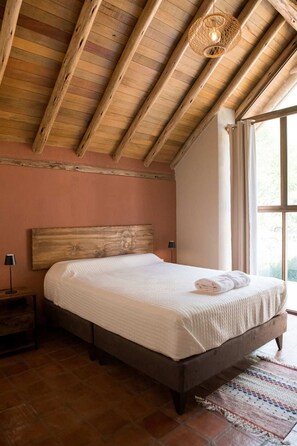 Un dormitorio con techo de madera, una cama grande con colcha blanca, una mesita de noche con lámpara y una ventana con cortinas.