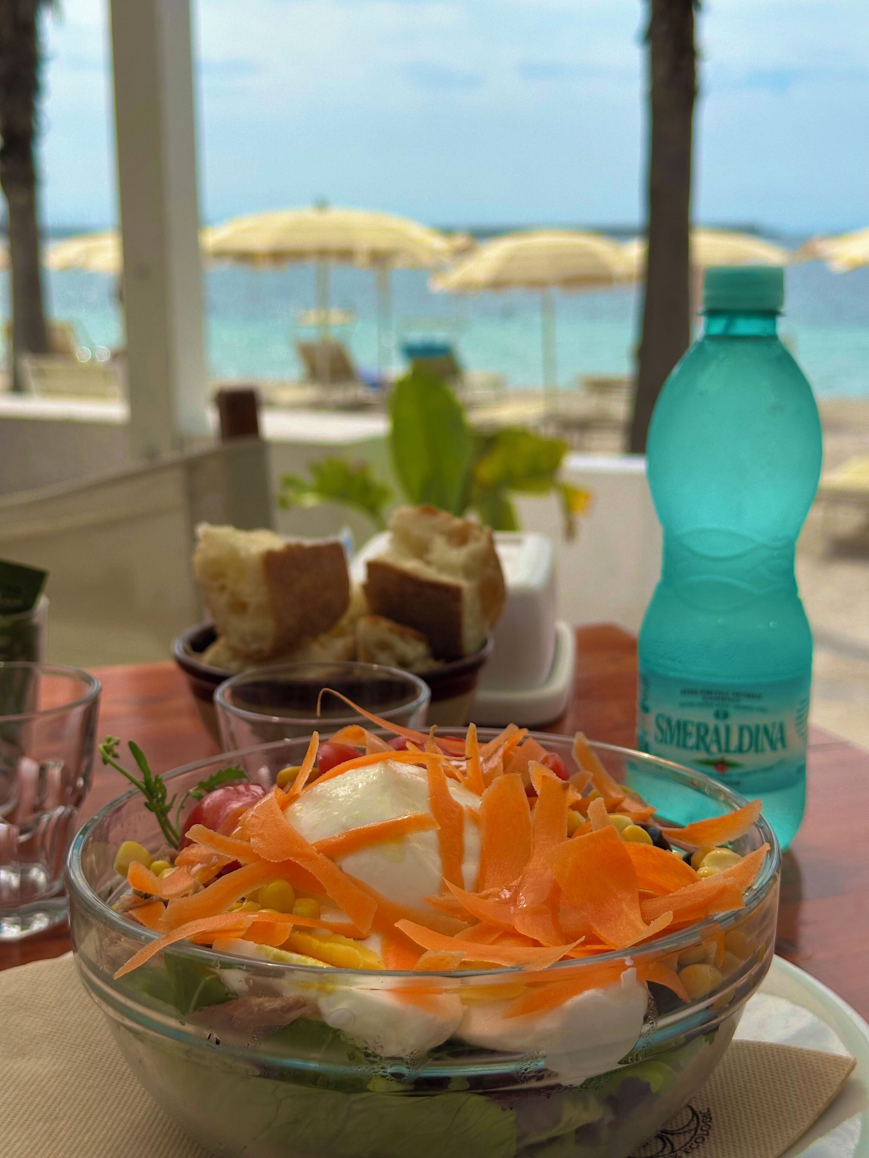 A glass bowl of salad with shredded carrots and a bottle of water on a table with a view of the beach and umbrellas in the background.