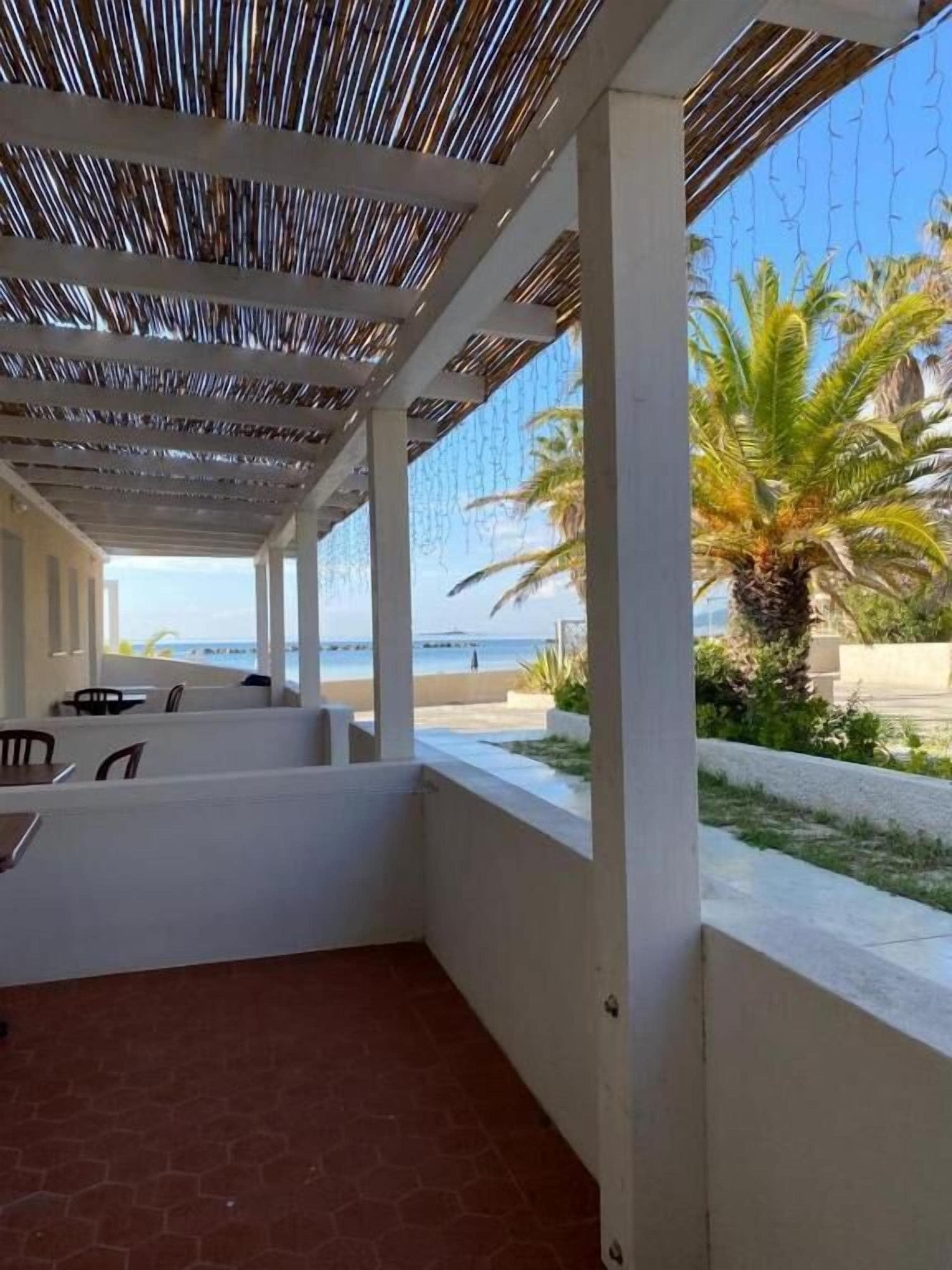 An outdoor patio area with a view of the beach, covered by a striped roof, and furnished with tables and chairs.