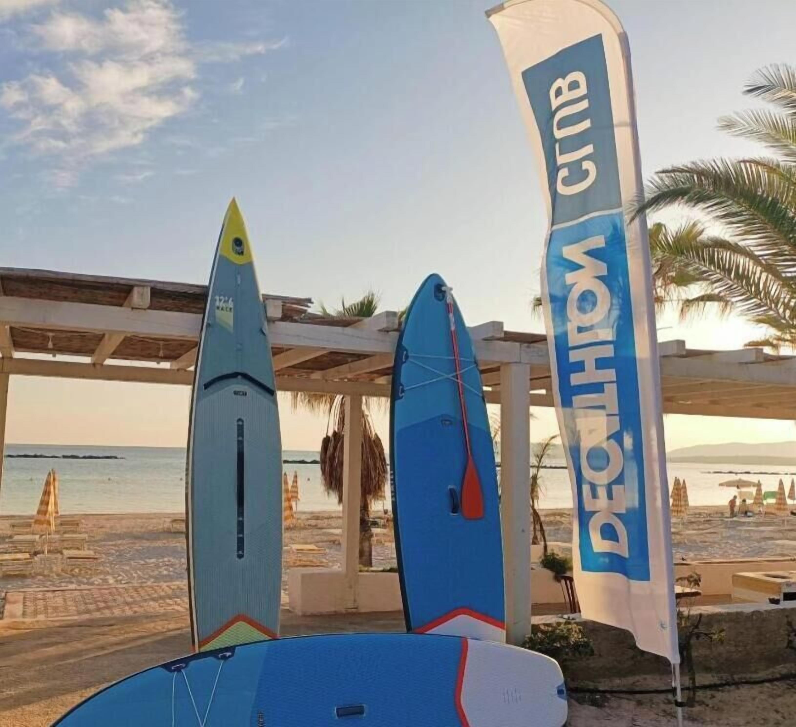 Three surfboards and a flag with the text "DECATHLON GTB" on a beach.