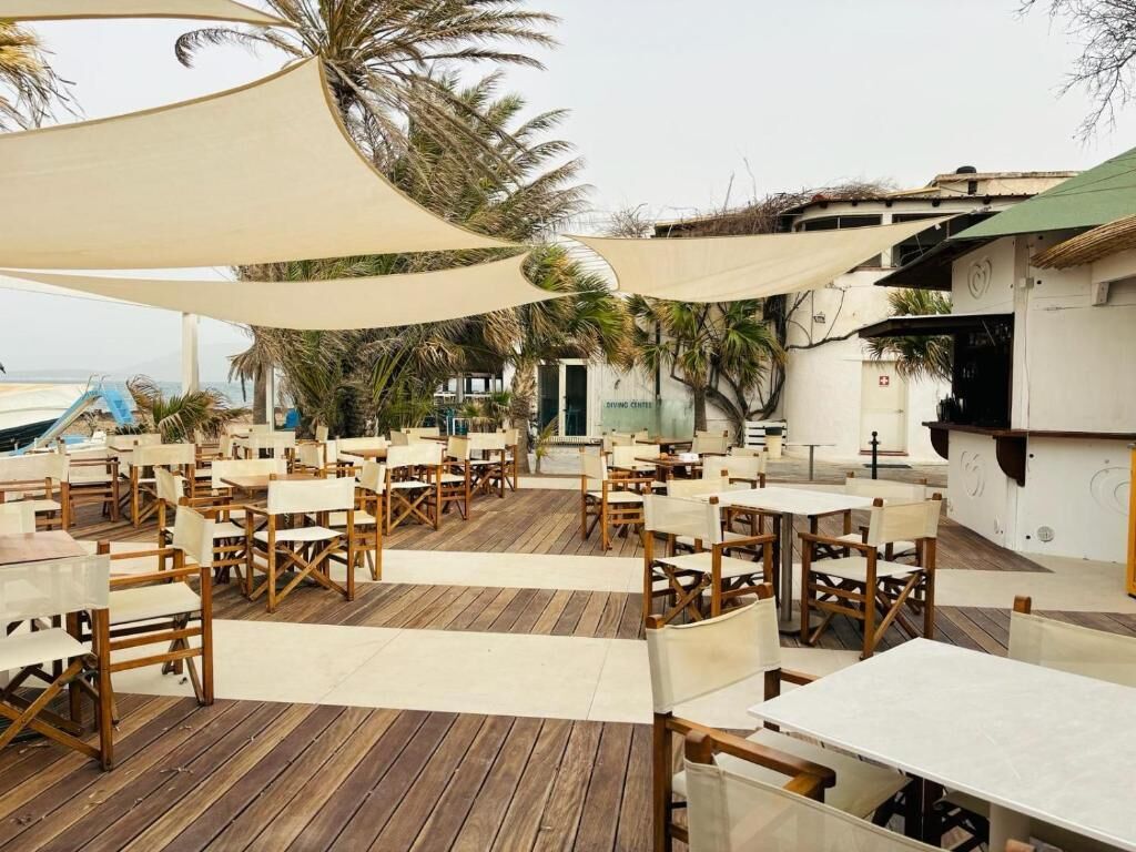 Un espace de restauration en plein air, avec des tables et des chaises en bois, à l’ombre de grands parasols blancs, donnant sur une plage.