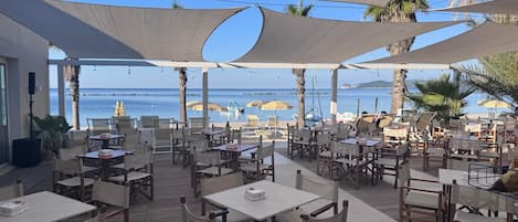 An outdoor dining area with tables and chairs, shaded by large canopies, overlooking a beach with boats and palm trees.