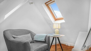 Ein modernes Schlafzimmer im Loft-Stil mit Dachfenster, einem grauen Sessel, einem kleinen Tisch mit Lampe und einem weißen Bettgestell.