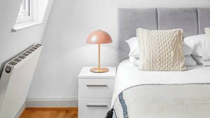 Ein Schlafzimmer mit schrägen Wänden, einem Bett mit grauem Kopfteil, weißer Bettwäsche, einer pinkfarbenen Lampe und zwei gerahmten abstrakten Kunstwerken an der Wand.