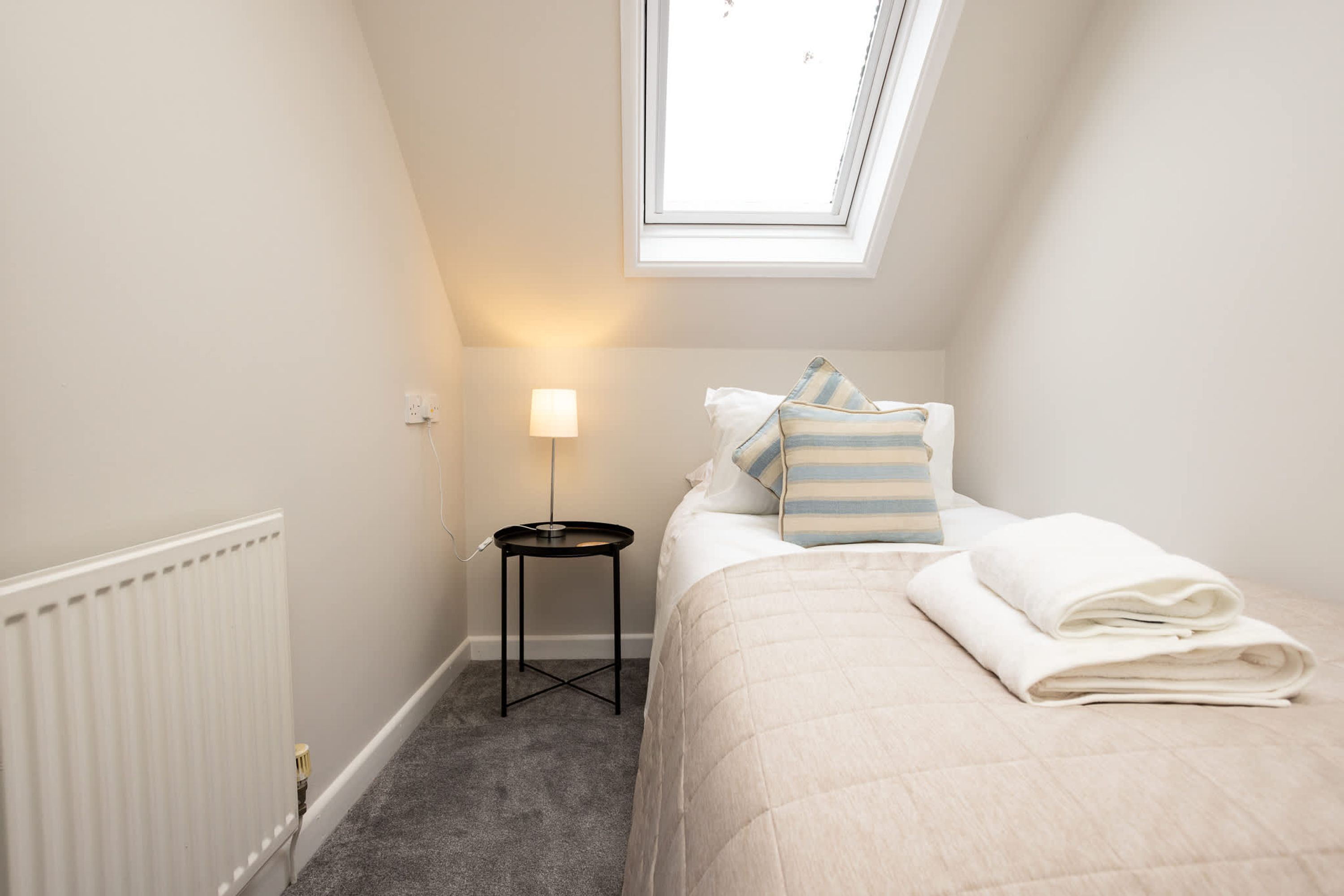 Een slaapkamer met dakraam, nachtkastje met lamp, bed met kussens en opgevouwen handdoek.