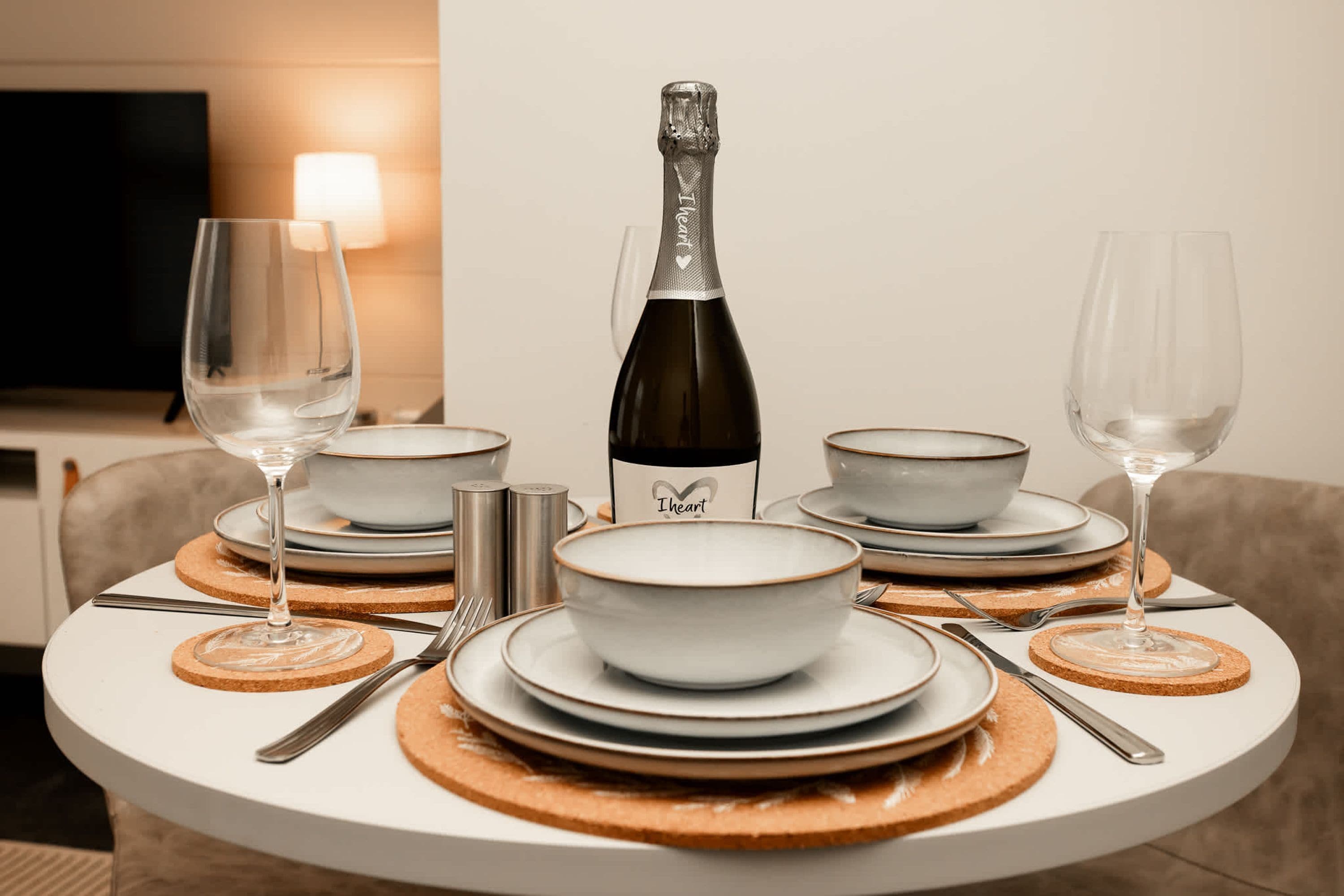 A dining table set with white plates, bowls, and cutlery, a bottle of champagne, and two glasses.