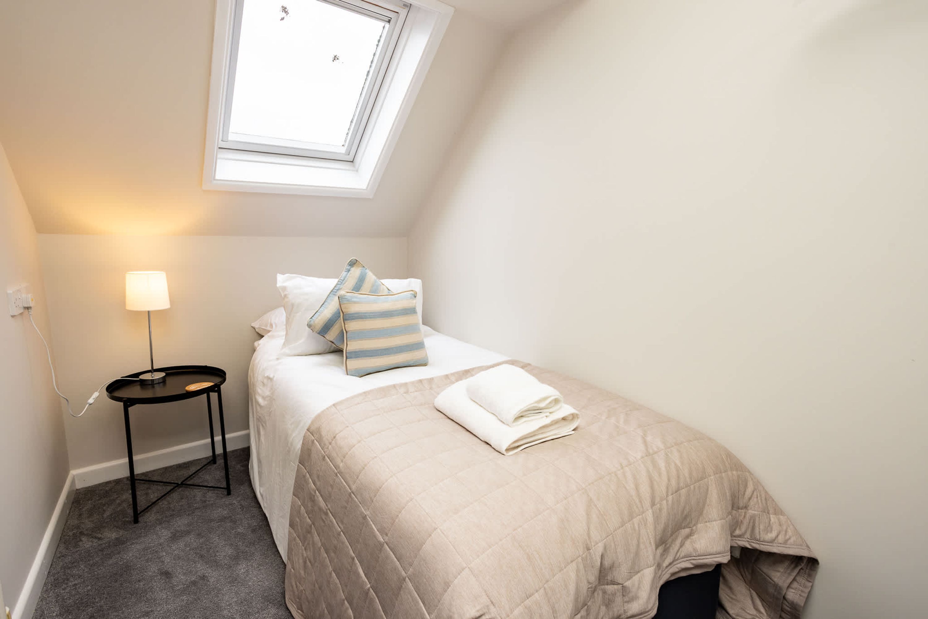 A small, neatly arranged bedroom with a single bed, a bedside table, a lamp, and a window.