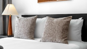 Una cama bien hecha con dos almohadas con textura, una lámpara de noche y una obra enmarcada en la pared.