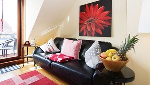 Un salon moderne avec un canapé en cuir noir, une table d’appoint en bois sur laquelle se trouve un panier de fruits, et un grand tableau représentant une fleur rouge accroché au mur.
