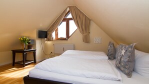 Une chambre avec un plafond en pente, un lit recouvert de draps blancs, une table de chevet ornée d’un vase de fleurs, une fenêtre avec des rideaux et un téléviseur fixé au mur.