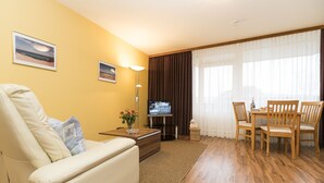 A living room with a sofa, a coffee table, a TV, a dining table with chairs, and a large window with curtains. - Hyggelige vacation apartment with swimming pool and sauna in Haus Ornum in Westerland (Westerland)