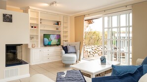 Een woonkamer met een open haard, een televisie, witte kasten, een comfortabele fauteuil en een groot raam met uitzicht op de omgeving.