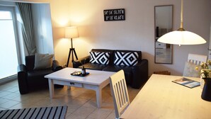 Ein modernes Wohnzimmer mit einer Couch, Couchtisch und Esstisch. Eine Lampe, ein Spiegel und ein an der Wand befestigtes Schild mit Koordinaten sind ebenfalls vorhanden.