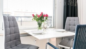 A dining area with a white table, four grey upholstered chairs, a vase with pink flowers, and a black curtain with a swirl pattern. - Modern vacation apartment with sauna, solarium and heated indoor pool in Westerland (Westerland)